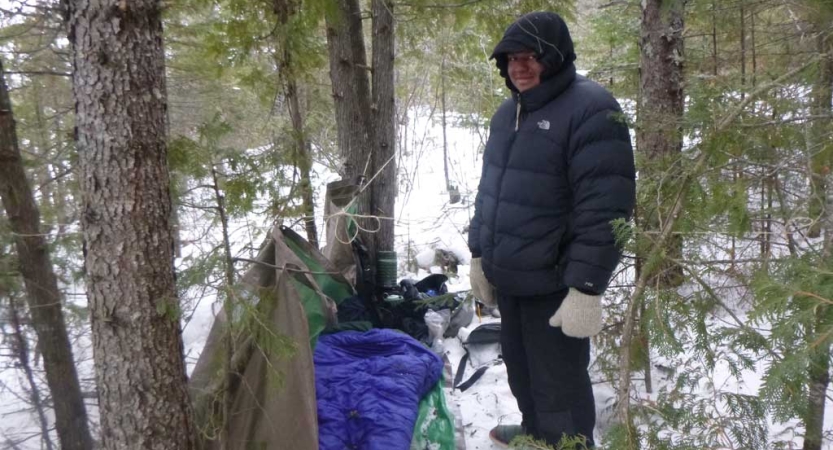 A person stands in a wooded, snowy area beside a tarp shelter. 